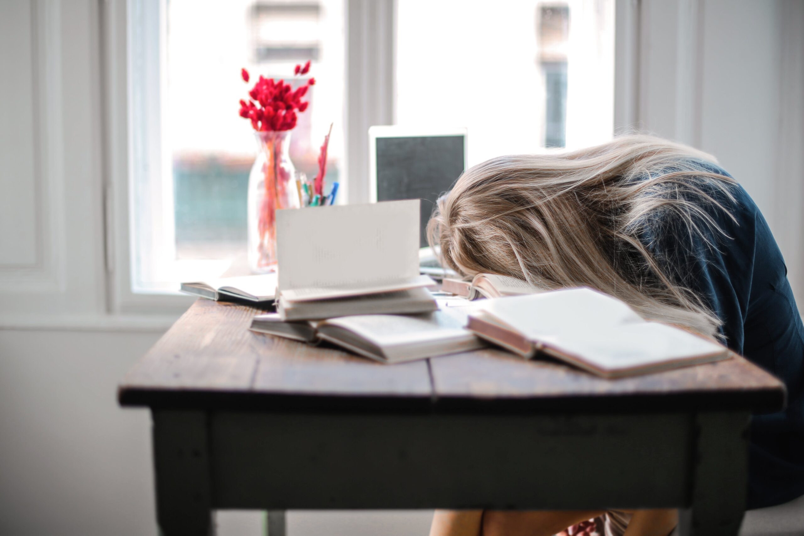 Blonde Frau liegt mit dem Kopf auf dem Tisch, neben ihr Bücher